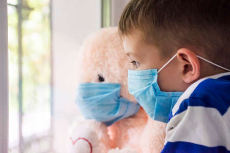 Sad child and his teddy bear both in protective medical masks looks out window. Virus protection, coronavirus pandemic. Coronavirus covid-2019.の写真素材
