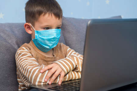 Boy studying at home in a medical mask. The child is remotely engaged using a laptop. Distance learning during quarantine concept.の写真素材