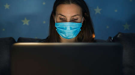 Young business woman in medical mask working from home with laptop in the evening during self-isolation and quarantine. Coronavirus outbreak and covid epidemic. Stay homeの写真素材