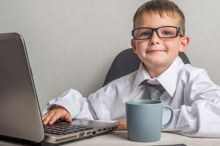 An adorable child is working on a laptop in suit and glasses. Little boss in the office. Children pretend to be adults.の写真素材