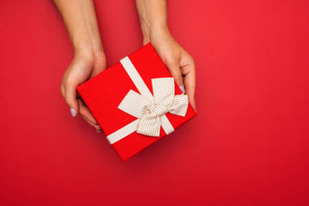 Female hands are holding present box or gift box with gold ribbon over red background. Top view.の写真素材