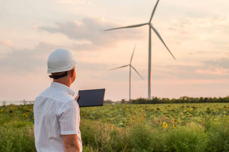 Engineer is checking energy production on wind turbine. Worker in windmills park in helmet and with tablet.の写真素材