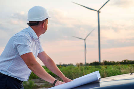 Young engineer working and holding blueprints at wind turbine farm power generator station. Engineer worker at wind turbine power station construction site.の写真素材
