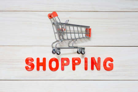 Empty shopping cart or market trolley on white wooden background with red inscription SHOPPING. Shopping cart and the word SHOPPING below. Concept of online shopping.の写真素材
