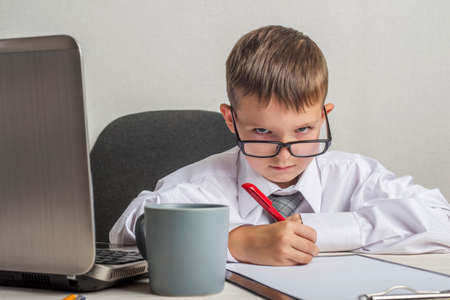 Serious child is writting on a clipboard in suit and glasses. Little boss in the office. Children pretend to be adults.の写真素材