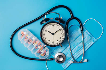 Composition with medical stethoscope, disposable face mask, alarm clock and pills on a blue background. Top view. Medical concept.の写真素材