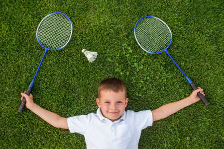 A smiling child holding two rackets in his hands lies on the grass next to the shuttlecock. Badminton game with a shuttlecock on the nature. Outdoor activity concept.の写真素材