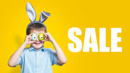 Shocked and surprised child in bunny ears and holding colorful easter eggs with painted big eyes on them in front of his eyes on a yellow background with text SALE.の写真素材