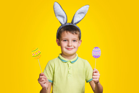 Cute boy with bunny ears holds colored eggs in his hands on yellow background. Happy child boy with easter eggs and bunny ears on yellow background.の写真素材