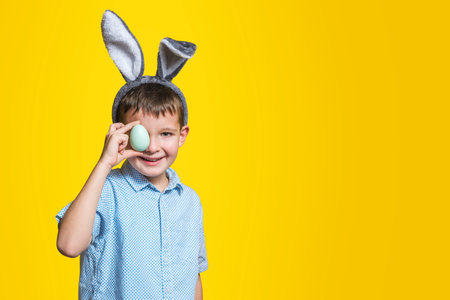 Happy child boy with bunny ears is covering his eye with Easter egg on yellow background. A kid with Easter egg on yellow background. Copy space.の写真素材