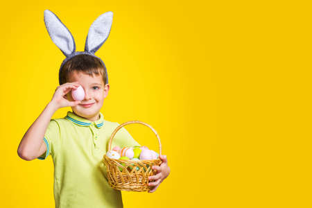 A happy smilling boy wearing Easter bunny ears holds a basket with painted eggs on a yellow background. Copy space. Easter day.の写真素材