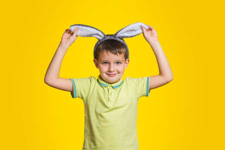 Little boy in t-shirt is touching his bunny ears and smiling while standing yellow background. Cute little child wearing bunny ears on Easter day.の写真素材