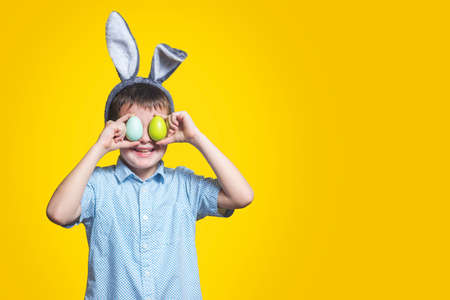 Cute boy with bunny ears keeping colored eggs near eyes on yellow background. Happy child boy with easter eggs and bunny ears on yellow background.の写真素材
