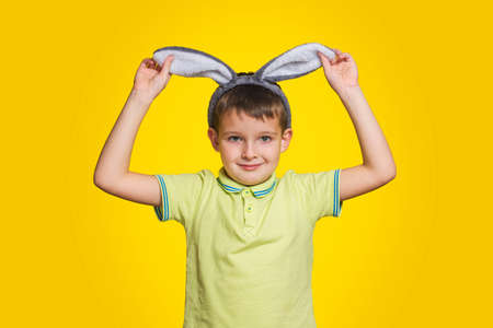 Little boy in t-shirt is touching his bunny ears and smiling while standing yellow background. Cute little child wearing bunny ears on Easter day.の写真素材