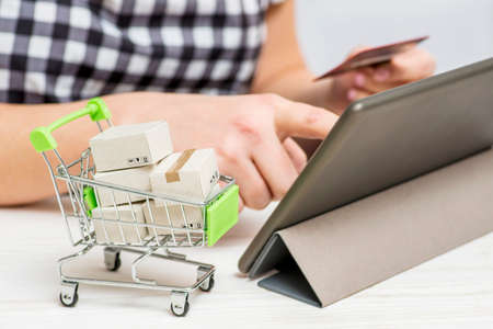 Paper boxes in trolley, womans hands holding credit card and using tablet to shop online. Online shopping conceptの写真素材