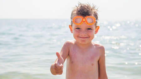 Happy little boy in glasses for swimming shows gesture - thumb up on the background of the sea. Travel and summer vacation concept.の写真素材