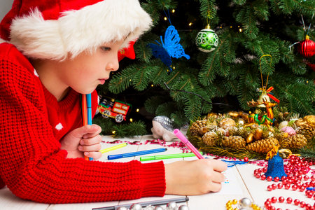 Cute boy in Santa hat writes a letter to Santa near the Christmas treeの写真素材