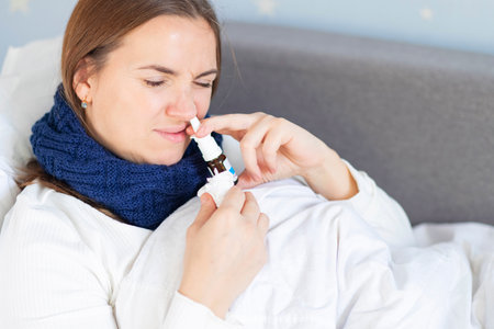 Young woman with bad cold lying in bed under blanket and using nasal dropsの写真素材