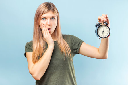 Shocked young blond woman with alarm clock on blue backgroundの写真素材