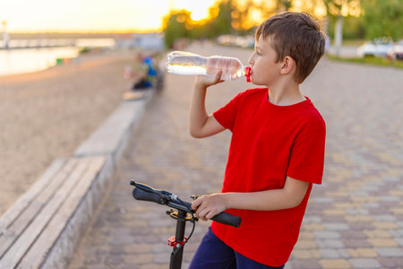 A boy drinks water from plastic bottle, standing with scooterの写真素材