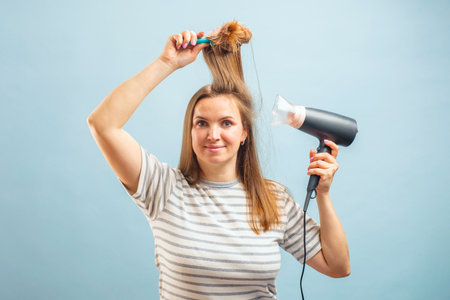 Young smiling woman using hair dryer on blue background. Hair care conceptの写真素材
