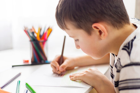 A child boy draws on white paper with colored pencils while sitting at a tableの写真素材