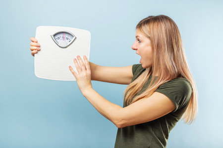 Young blonde woman looking at weight scale in shock on blue backgroundの写真素材