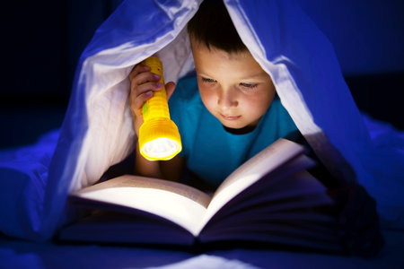 A young boy reading a book under the covers with a flashlight at dark night timeの写真素材
