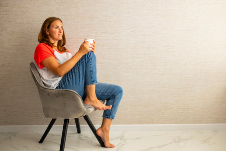 Young beautiful woman in casual wear sitting in armchair with cup of hot coffeeの写真素材