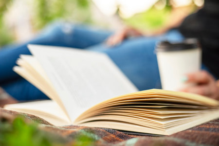 A woman in jeans with a book and a cup of coffee sits on a blanket in the parkの写真素材