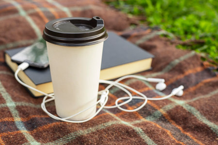 Cup of coffee, open book and smartphone with headphones on blanket in the parkの写真素材