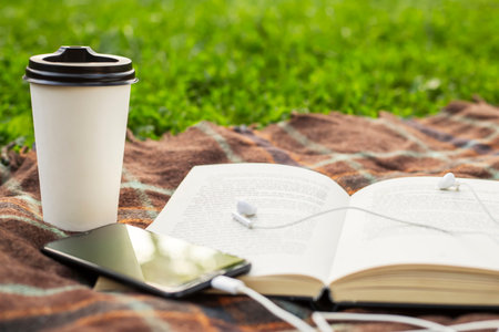Cup of coffee, open book and smartphone with headphones on blanket in the parkの写真素材