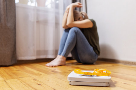 Upset and sad woman sitting on wooden floor near white scale with measuring tapeの写真素材