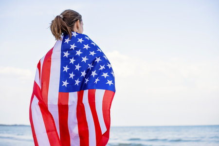 Back view of young woman holding national American flag on a ocean beachの写真素材