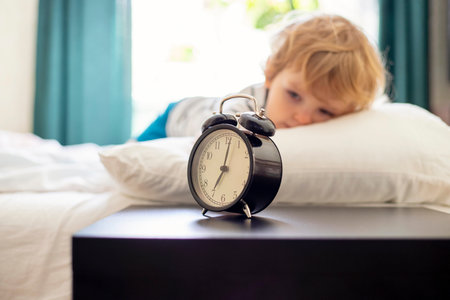 A sleepy little child lies on the bed and watches the ringing alarm clock