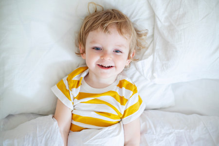 Top view of cheerful little boy in white bed early in the morningの写真素材