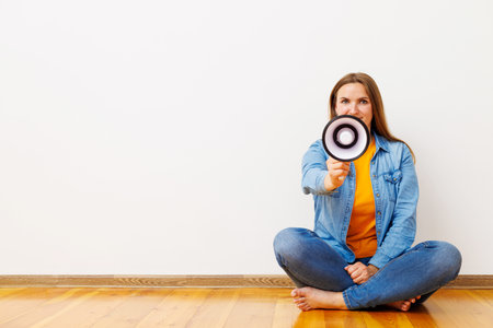 Young woman screaming on megaphone sitting on floor in front of white wallの写真素材