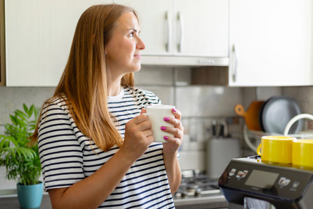 Contemplative woman holding a coffee mug in a sunlit kitchenの写真素材