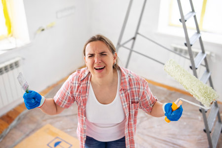 Frustrated woman with paintbrush and roller during home renovationの写真素材