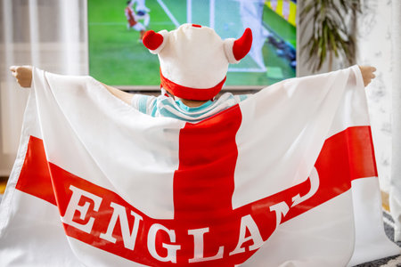 A boy holding England flag and watching soccer game on TV at homeの写真素材