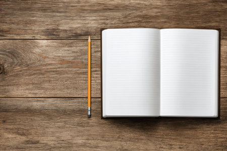 Overhead view of an open notebook and pencil on a wooden desk. The composition includes empty space on the wood grain surface, ideal for adding quotes or titles.の素材