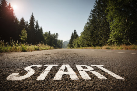 A long, empty road lined with green trees features the word "START" painted on the asphalt. Captured on a bright sunny day, the image conveys a sense of optimism and new beginnings.の素材