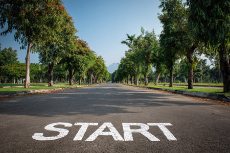 A long, empty road lined with lush green trees under a bright sunny sky. The word "START" is painted on the asphalt, symbolizing new beginnings and optimism.の素材