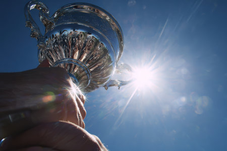 A dramatic close-up of hands gripping a heavy silver trophy, captured from below with a bright blue sky and lens flare in the background, symbolizing victory and achievement.の素材