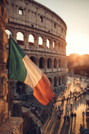 The Italian flag waves gracefully over the Colosseum at sunset, casting a warm glow on ancient stones. A cinematic Roman atmosphere with a tourist-filled plaza in soft focus.の素材