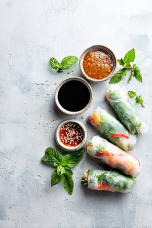 A clean and minimalistic food layout featuring vibrant rice paper spring rolls accompanied by three dipping sauces, elegantly arranged on a white tile background for modern food styling.の素材