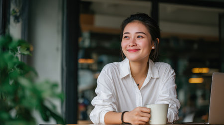 Young Asian woman in a white shirt sits at a desk with a coffee mug and laptop, smiling thoughtfully in a cozy modern office environment, embodying a lifestyle corporate vibe.の素材