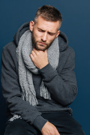 A man wearing a hoodie and scarf sits against a dark blue background, holding his throat with a painful expression. Ideal for advertisements related to sore throat remedies.の素材