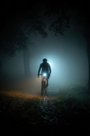 A cyclist in full gear rides through a dark park trail, illuminated only by bike lights and a headlamp, creating a striking silhouette framed by faint fog.の素材