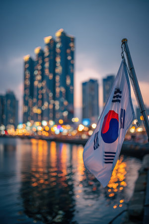 The South Korean flag flutters in Busan harbor at dusk, with cranes and skyscrapers in the background. Modern neon reflections create a vibrant, futuristic atmosphere in a wide frame.の素材